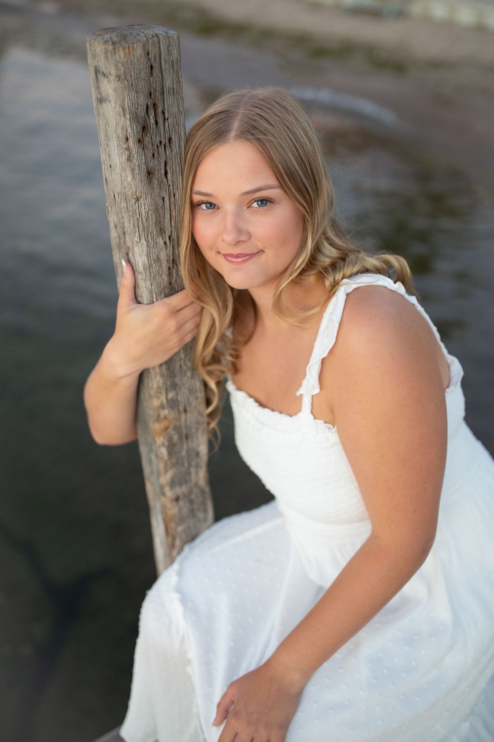 Senior portrait of a girl in a white dress posing near wooden post on a dock at West Lake Okoboji.