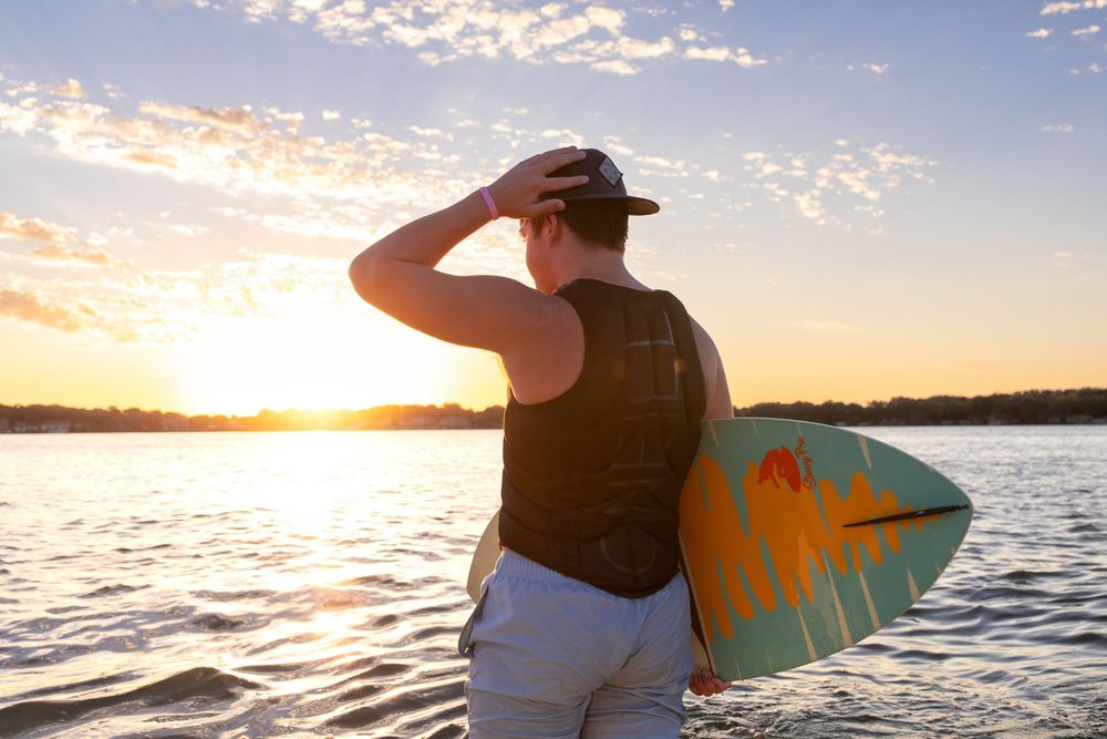 Surfer holding a teal surfboard watches a vibrant orange sunset over calm lake waters at West Okoboji Lake.