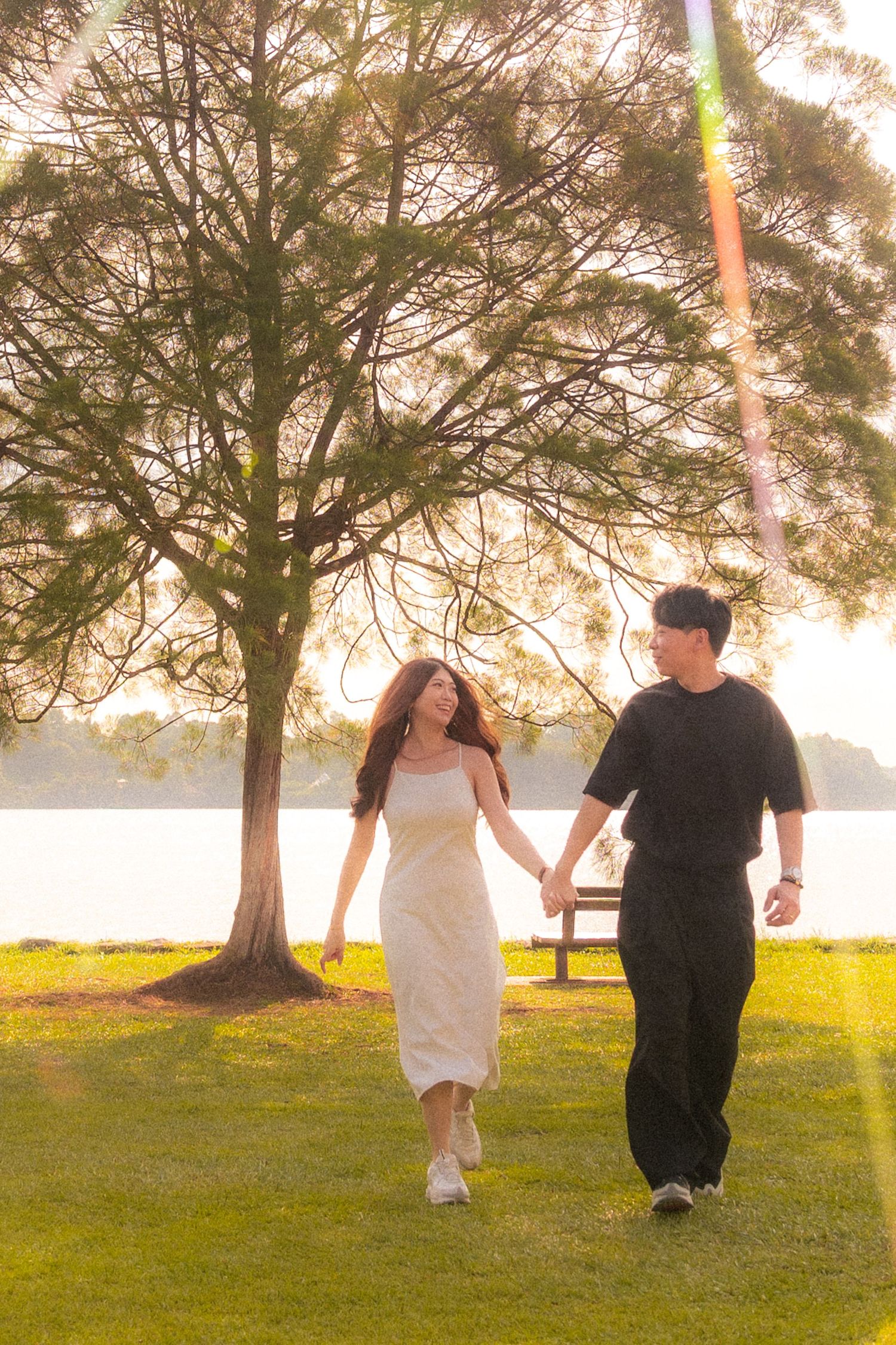 A couple walks hand in hand through a sunlit park with a lake and pine trees in the background during golden hour.
