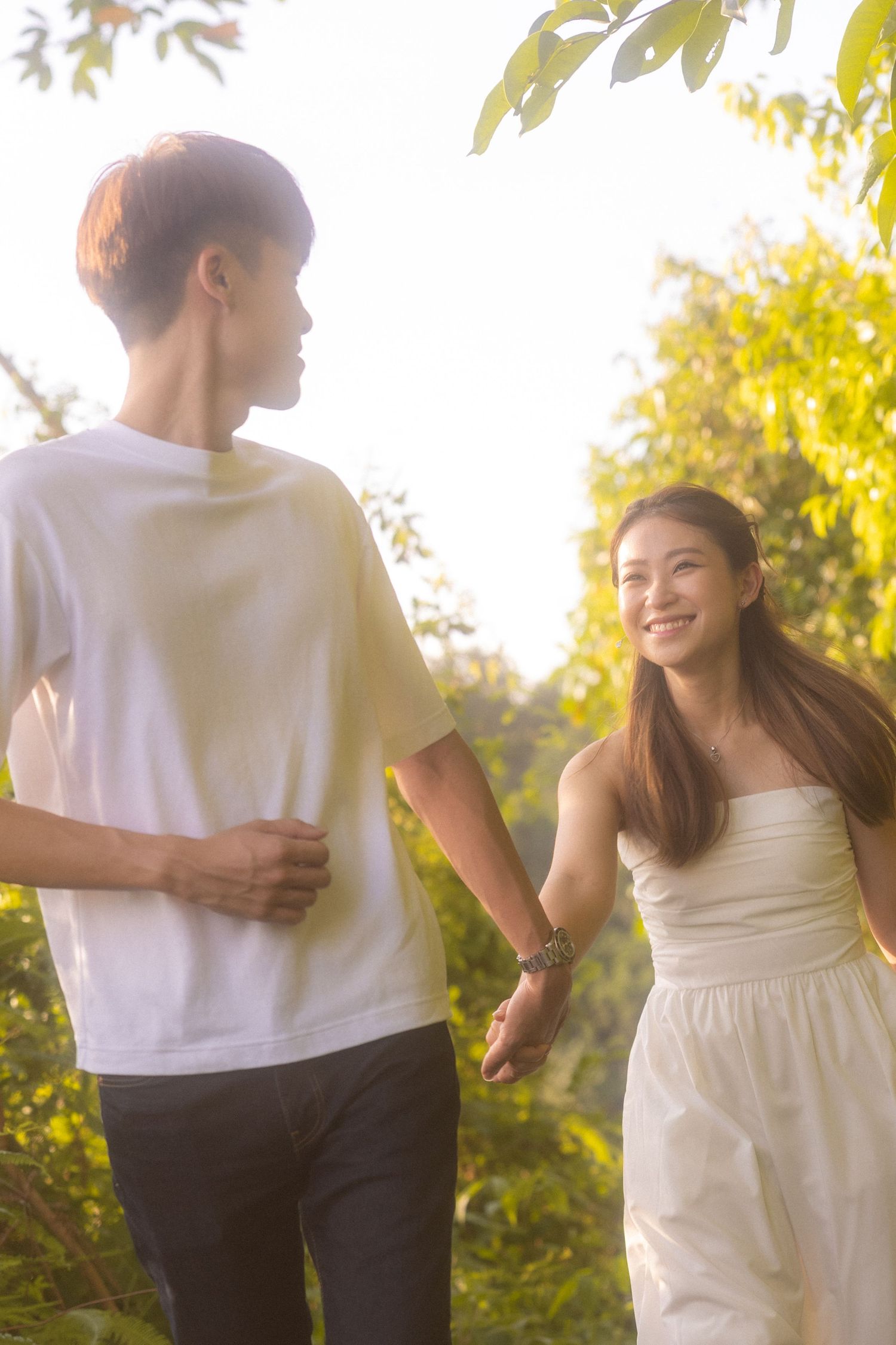 Young couple holding hands while walking together in a sunlit garden with golden foliage in the background.
