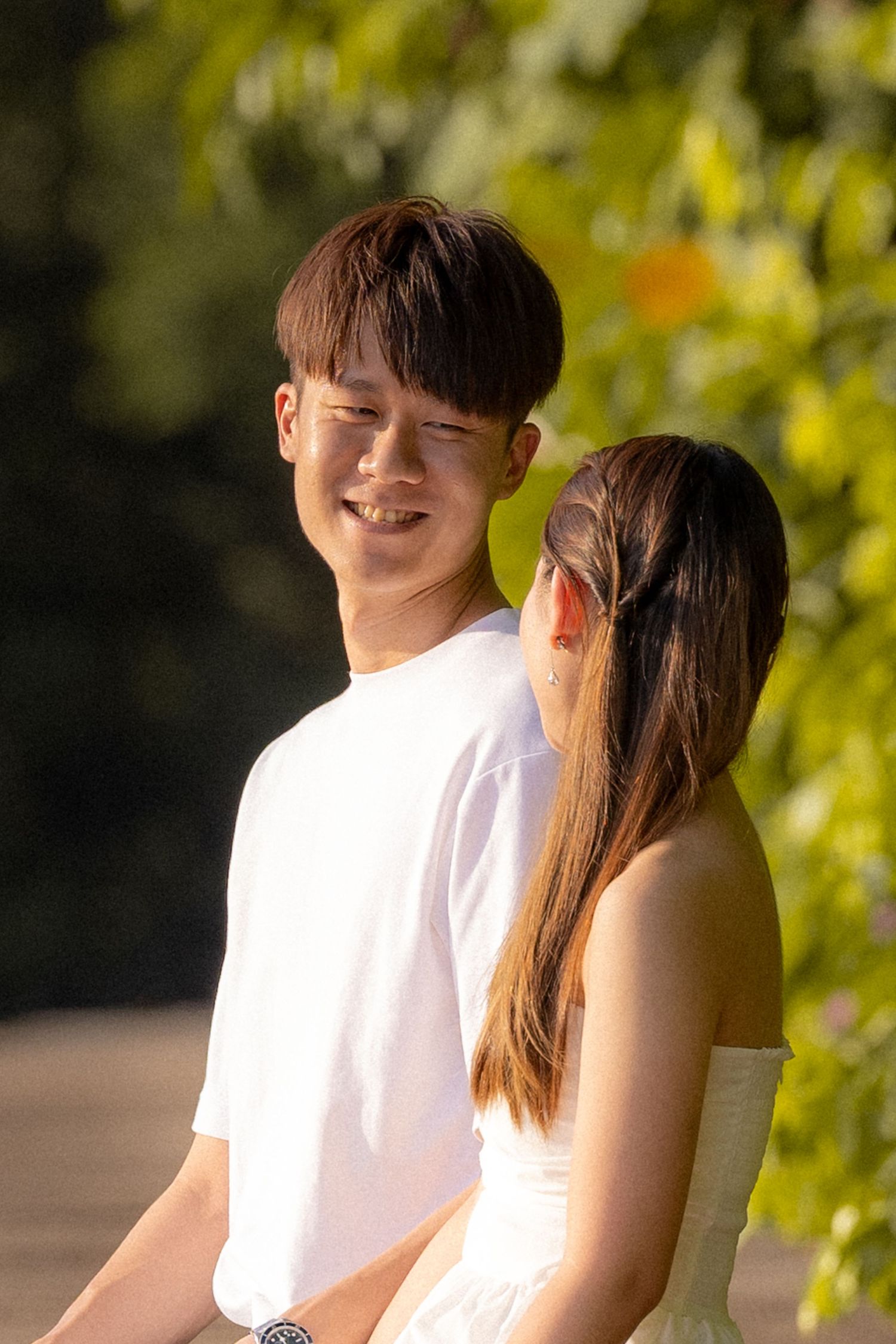 A couple dressed in white clothing sits together outdoors in natural sunlight with green foliage in the background.