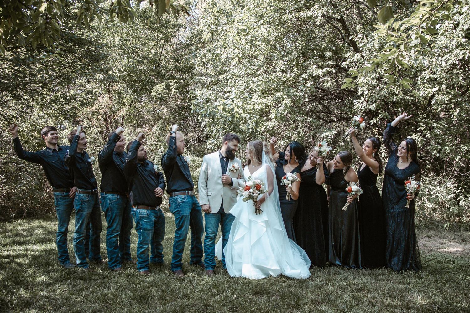 Wedding party in blue and black attire poses playfully in outdoor garden setting.
