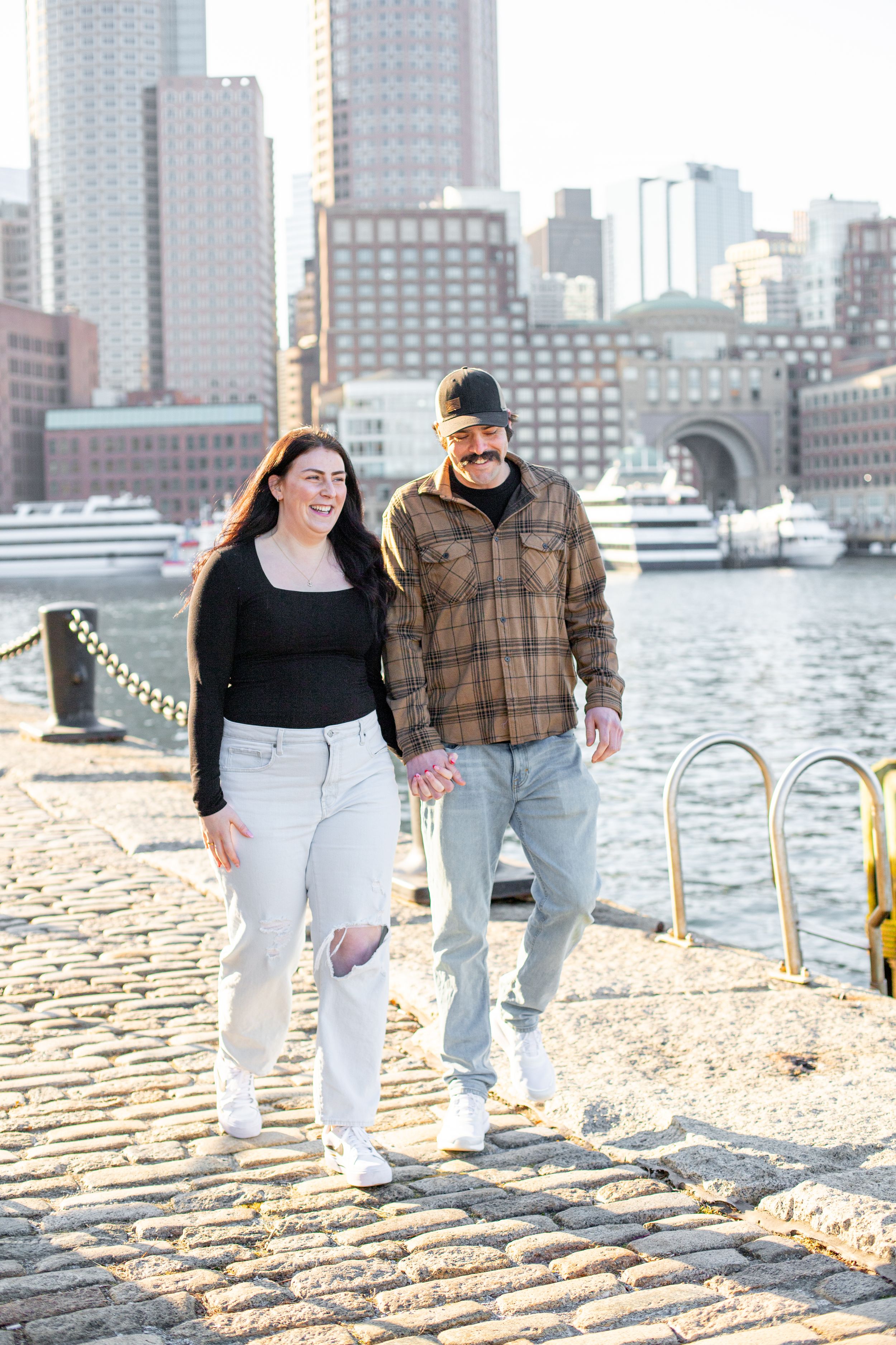 photoshoot in boston seaport; couple walks along fan pier park with boston in the background