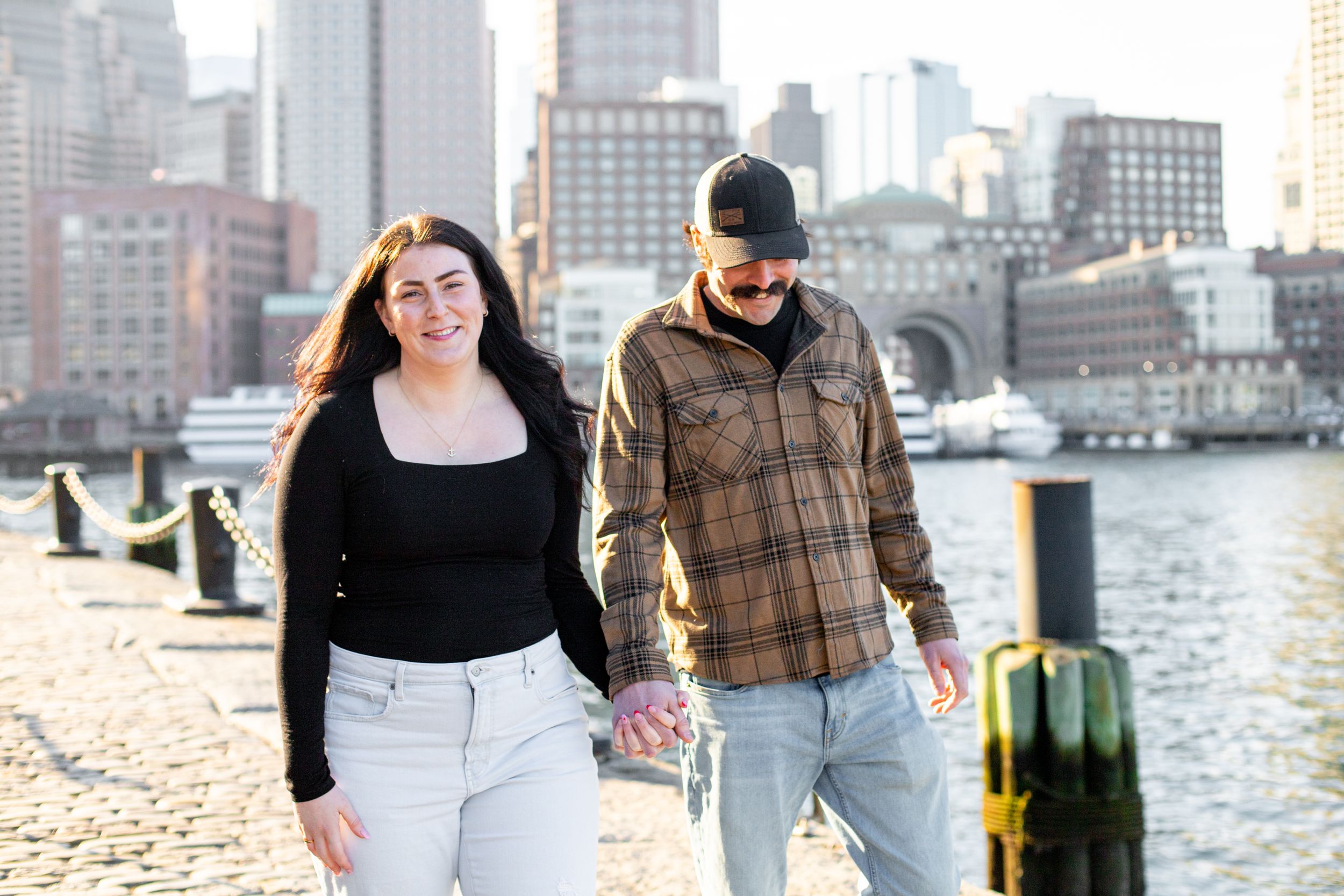 photoshoot in boston seaport; couple walks along fan pier park with boston in the background