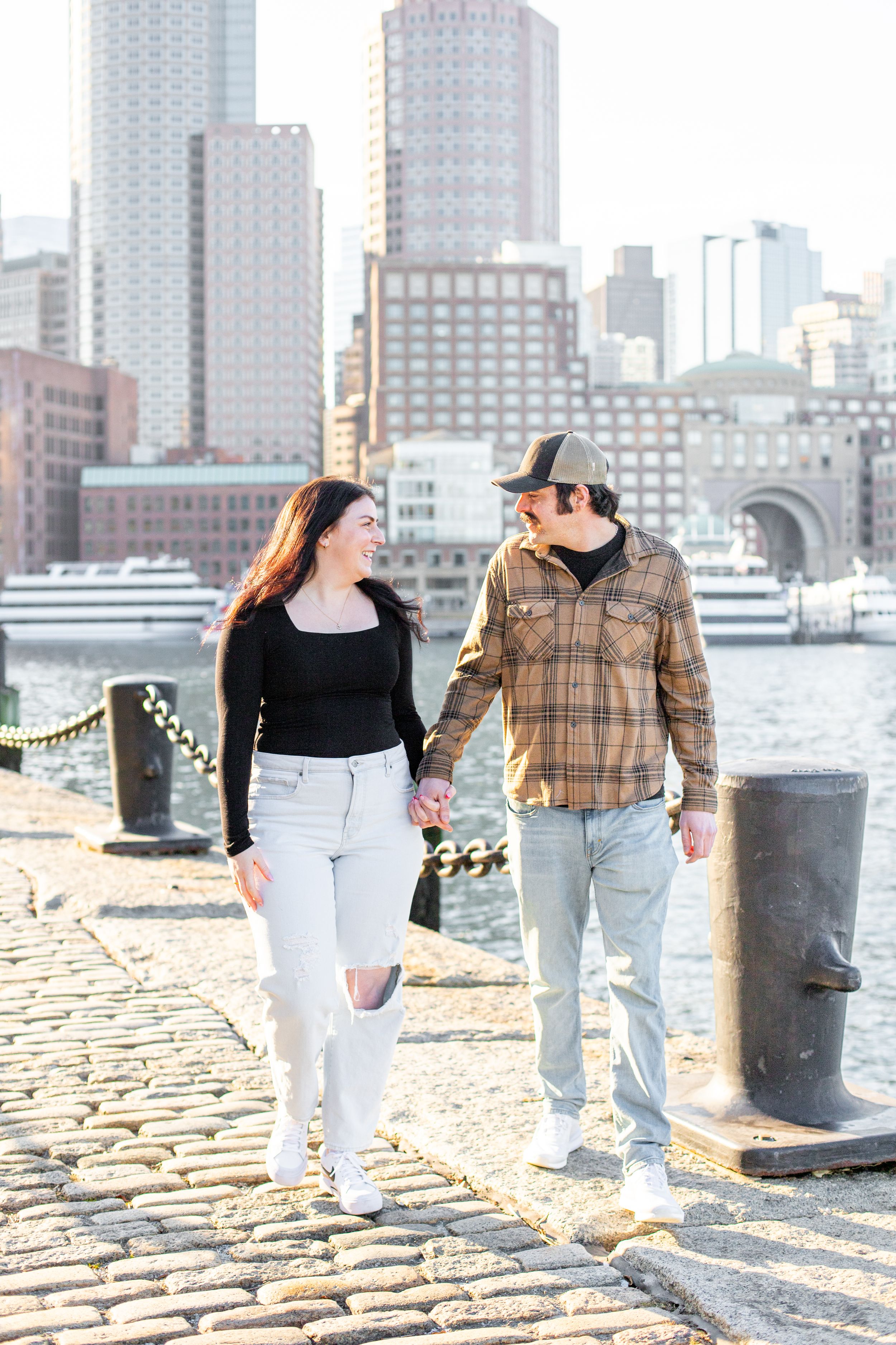 photoshoot in boston seaport; couple walks along fan pier park with boston in the background