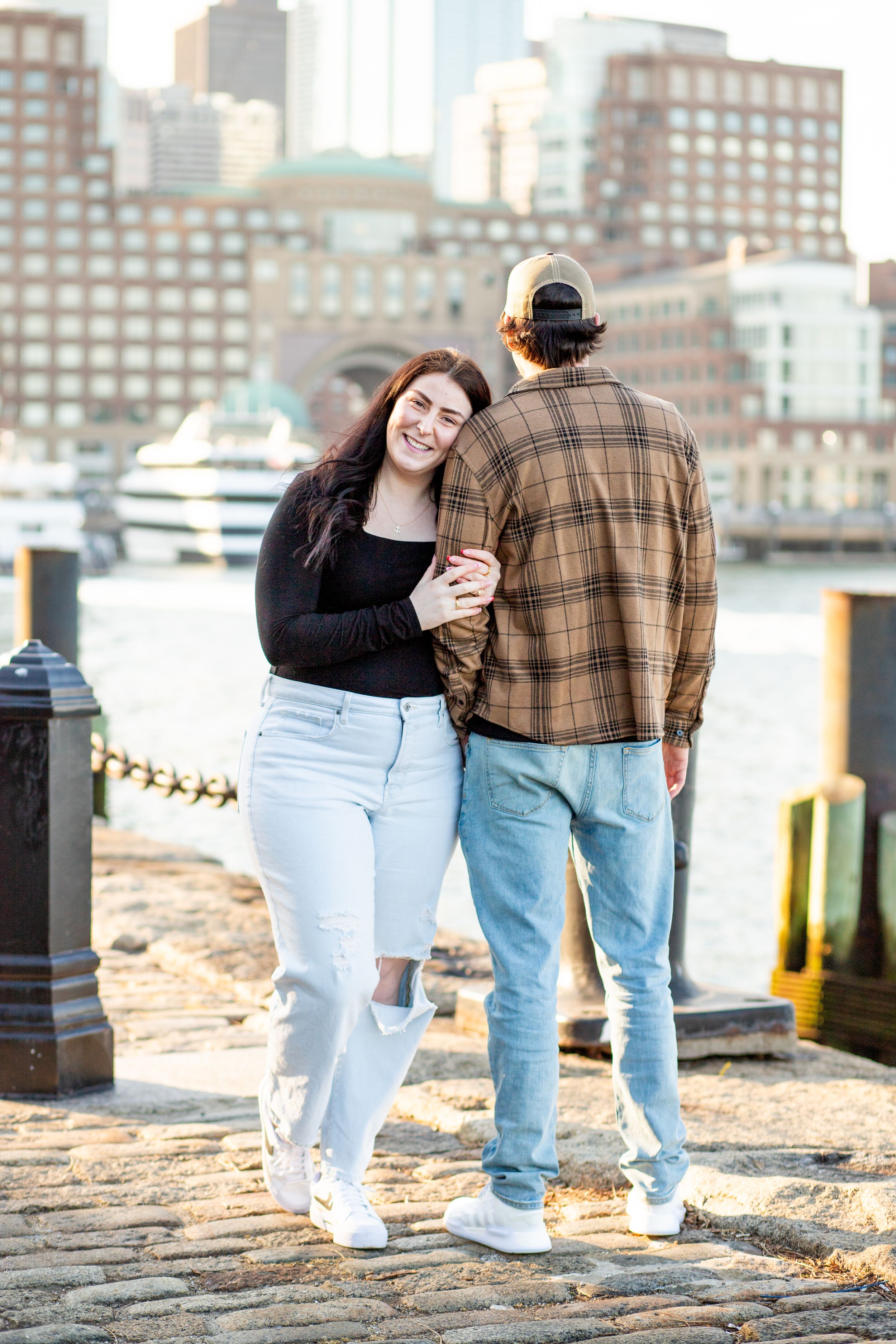 photoshoot in boston seaport; couple walks along fan pier park with boston in the background at golden hour