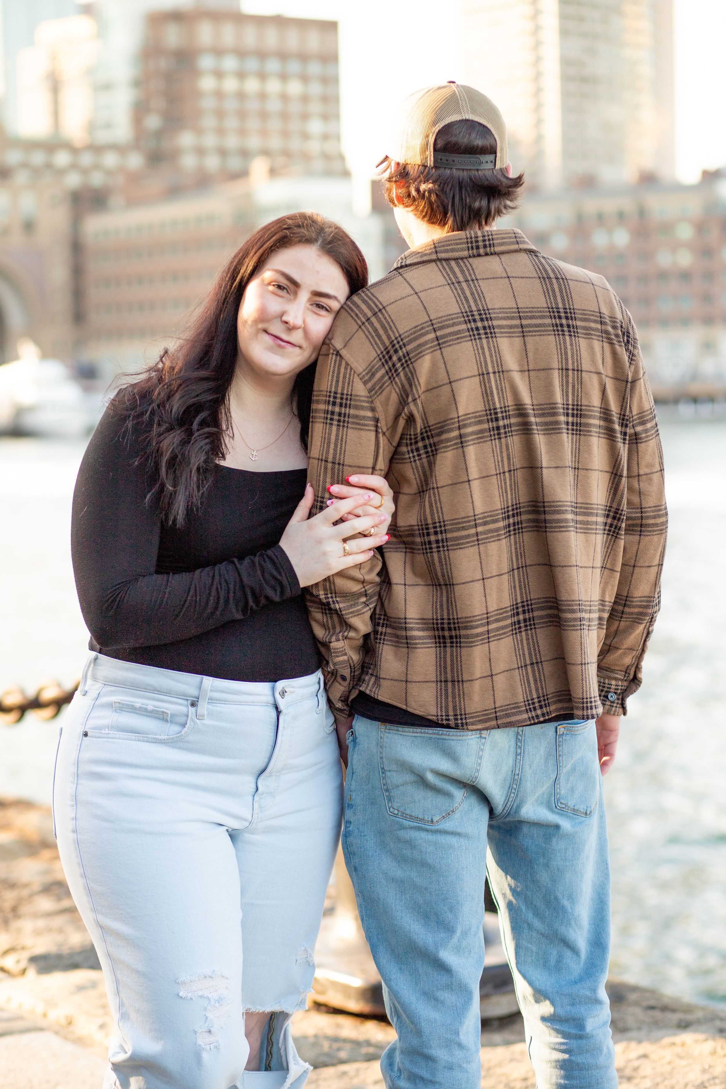 photoshoot in boston seaport; couple walks along fan pier park with boston in the background at golden hour