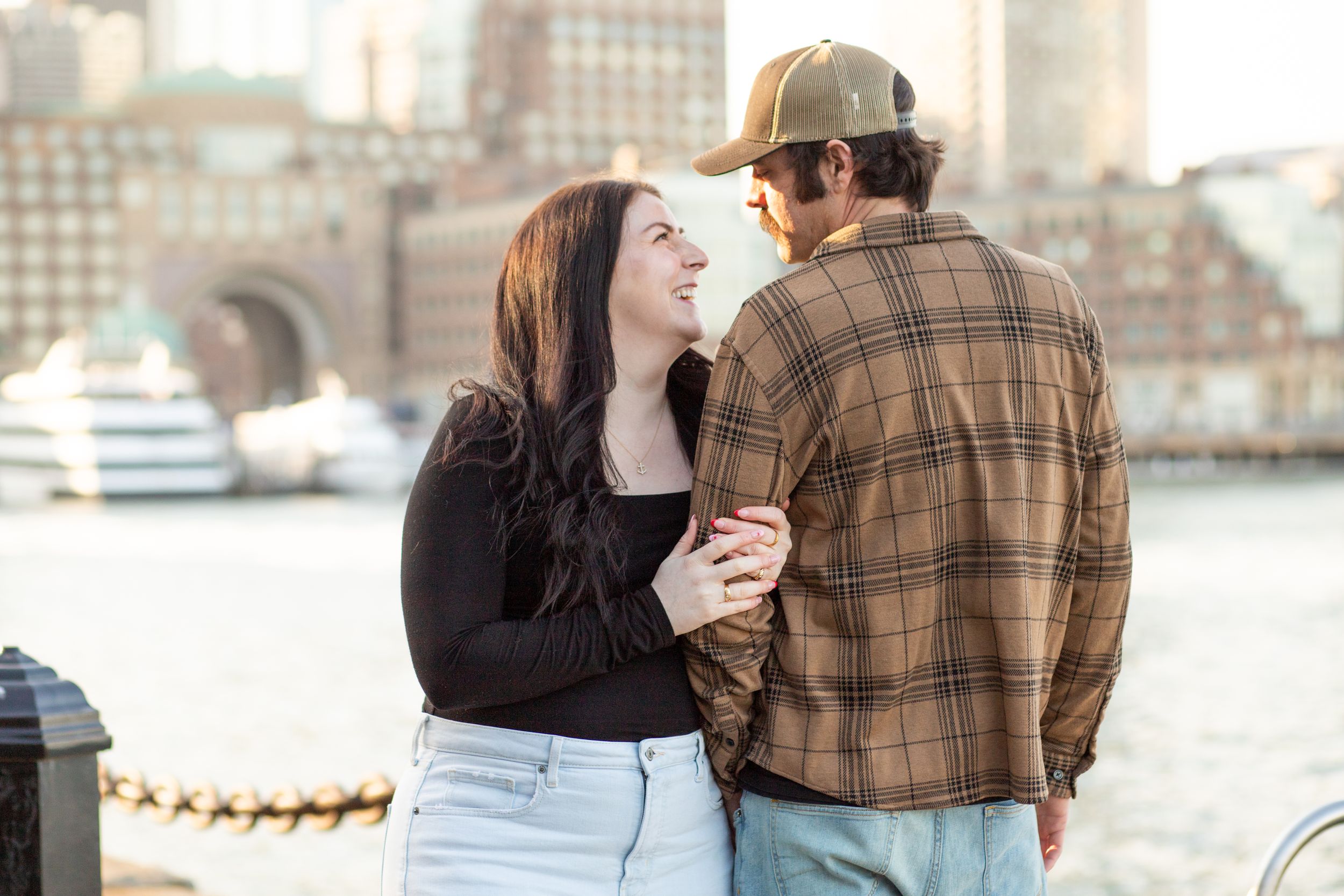 photoshoot in boston seaport; couple walks along fan pier park with boston in the background at golden hour