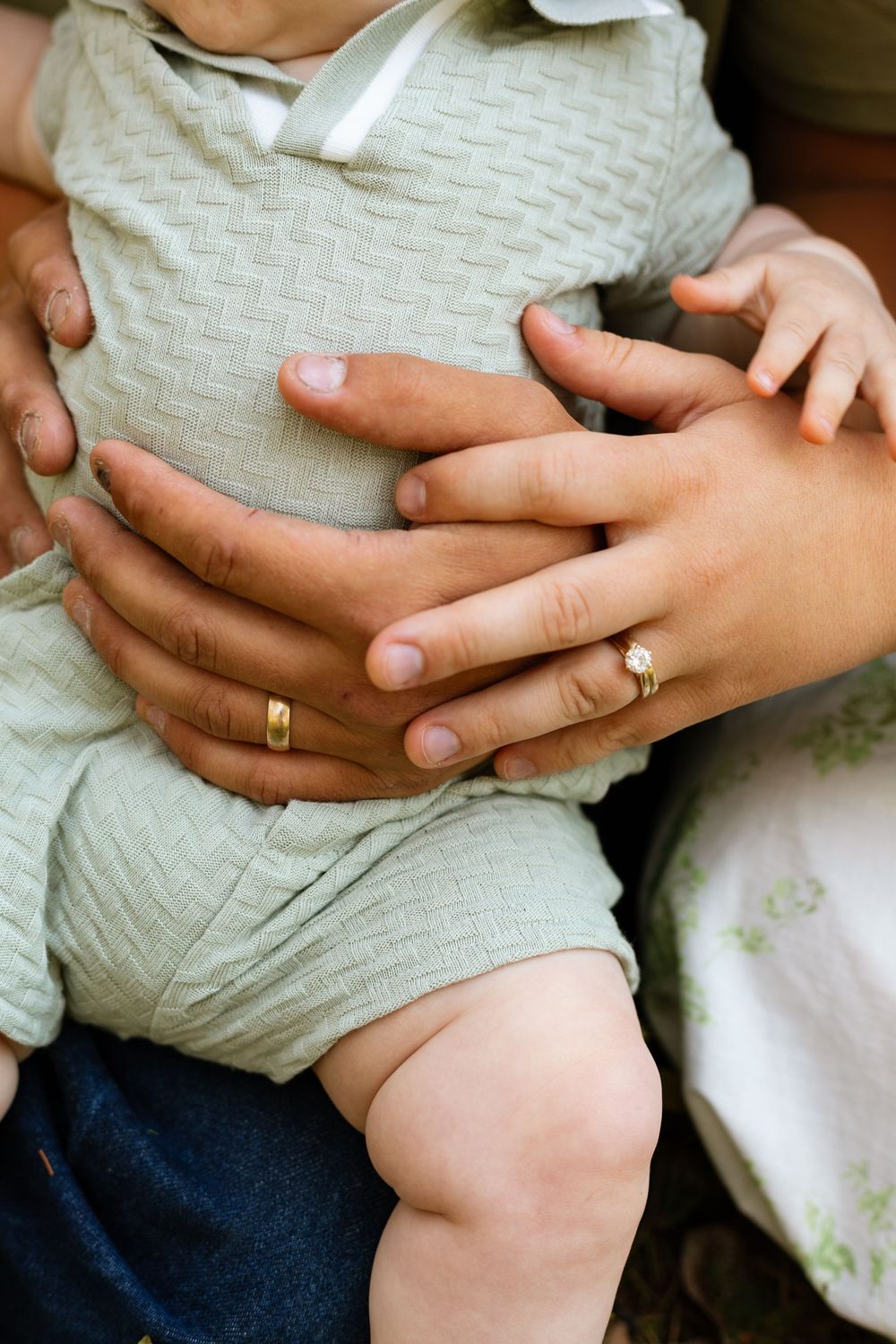Two parents wearing wedding rings lovingly hold their baby in a green knit onesie.