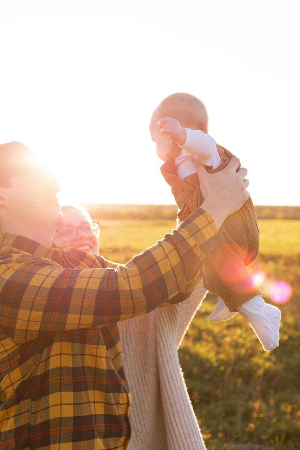 Parent lifts small baby in golden sunset light while wearing plaid shirt in autumn field.