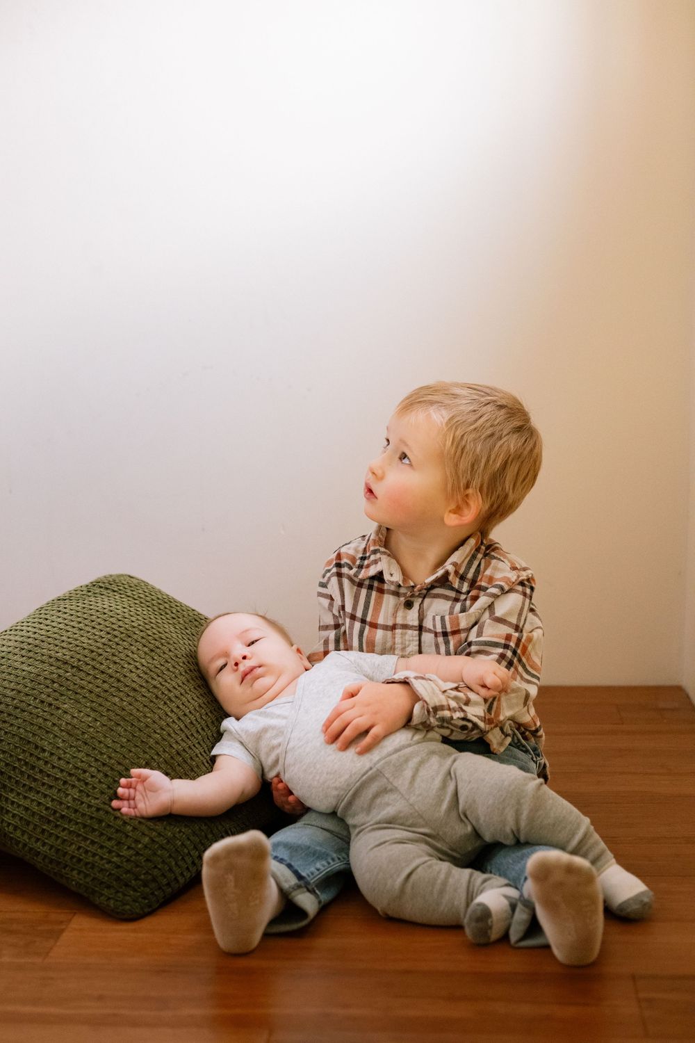Two young children lie on hardwood floor with green pillow while looking up at ceiling in playful poses.
