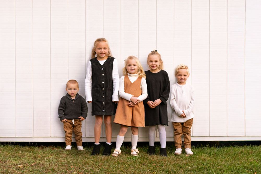 Children sit together against a white wall wearing coordinated fall outfits in brown, black and cream colors.