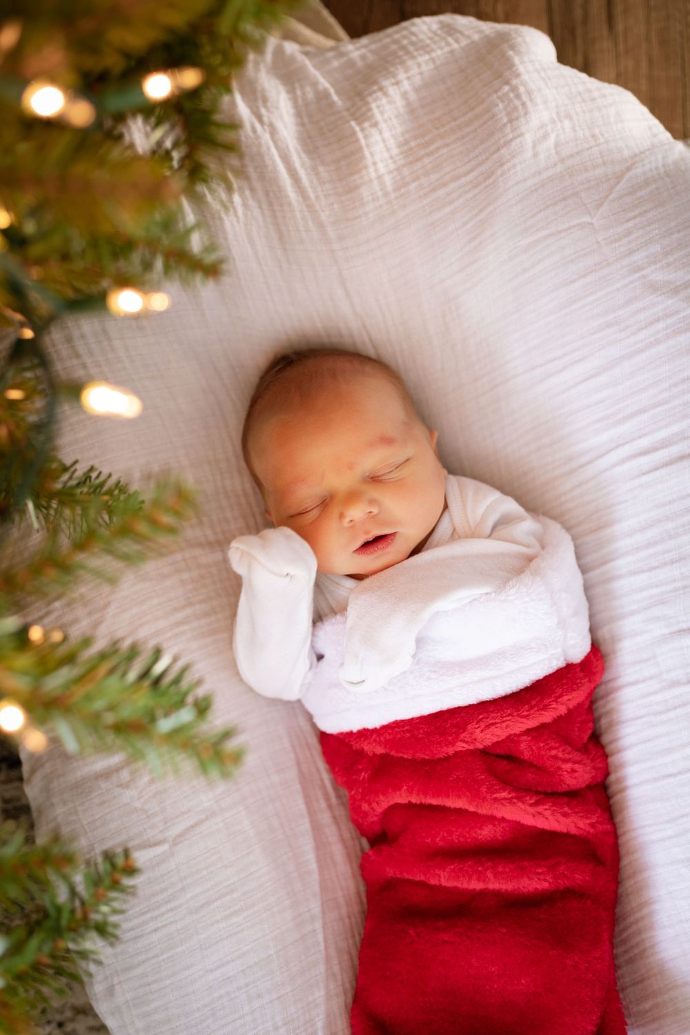 A sleeping baby wrapped in a stocking next to Christmas tree lights.