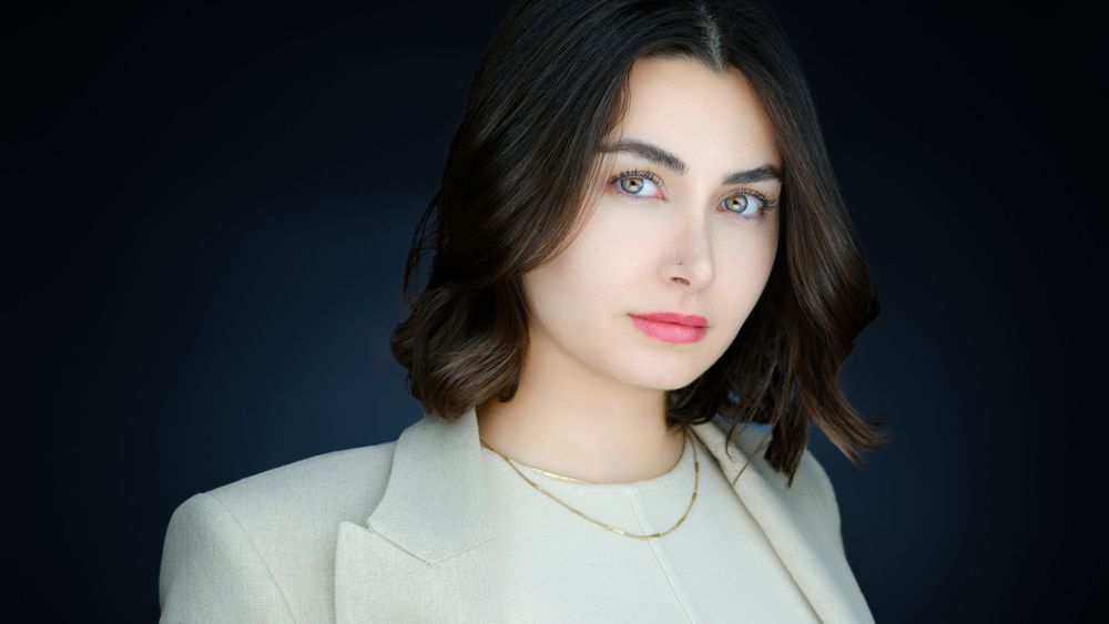 Professional headshot of a young woman with dark hair wearing a white blazer and gold necklace.