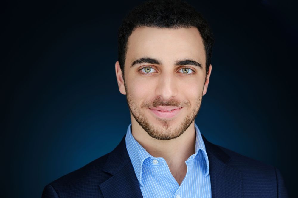 Professional headshot of a smiling young man with light eyes wearing a blue suit and light blue shirt.