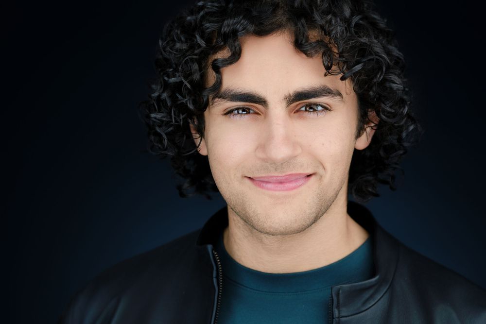 Professional headshot of young man with curly dark hair in leather jacket.