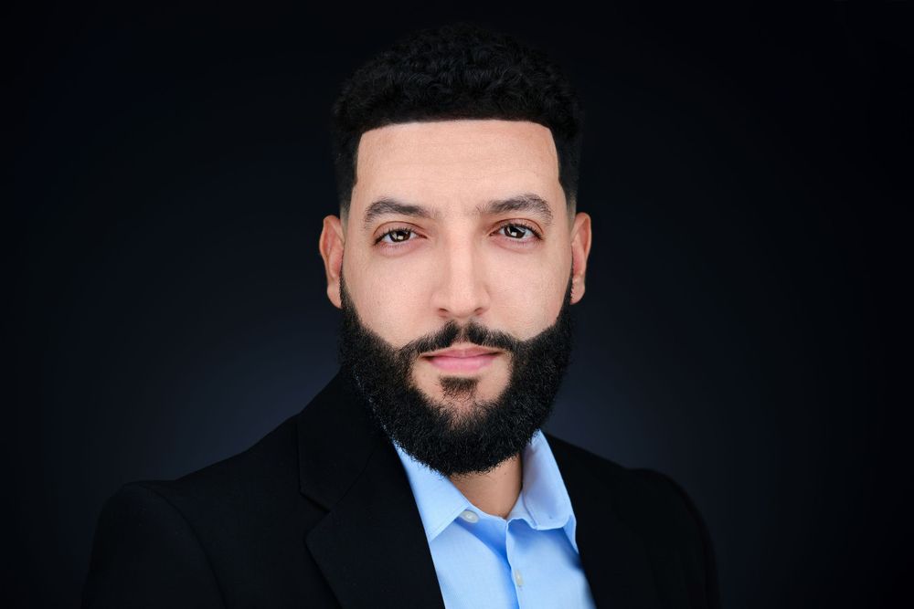 Professional headshot of bearded man in black blazer and blue shirt on dark background.
