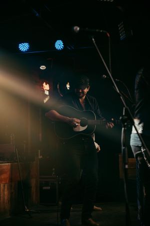 Musician performs on dark stage with acoustic guitar under dramatic lighting and blue spotlights above.