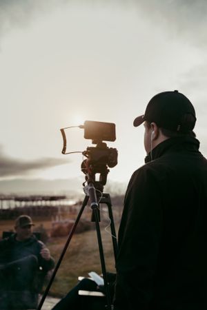 A photographer in a dark coat operates a camera on a tripod during sunset, capturing a moody outdoor scene.