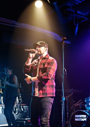 Singer in a red plaid shirt performs passionately on stage with dramatic lighting.