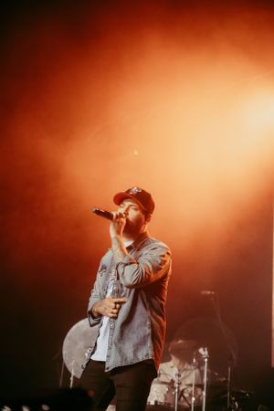 A performer in a baseball cap sings on stage with dramatic orange lighting during a concert performance.