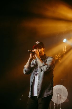 A performer in a denim jacket sings on stage with dramatic orange lighting and a drum kit visible in the background.