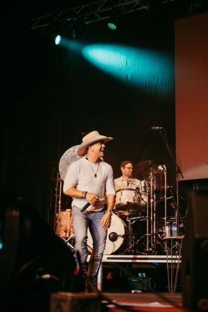 Country musician performs on stage wearing a cowboy hat while a drummer plays in the background under blue lighting.