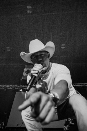 Black and white photo of a performer in a white cowboy hat holding a microphone on stage during a concert performance.