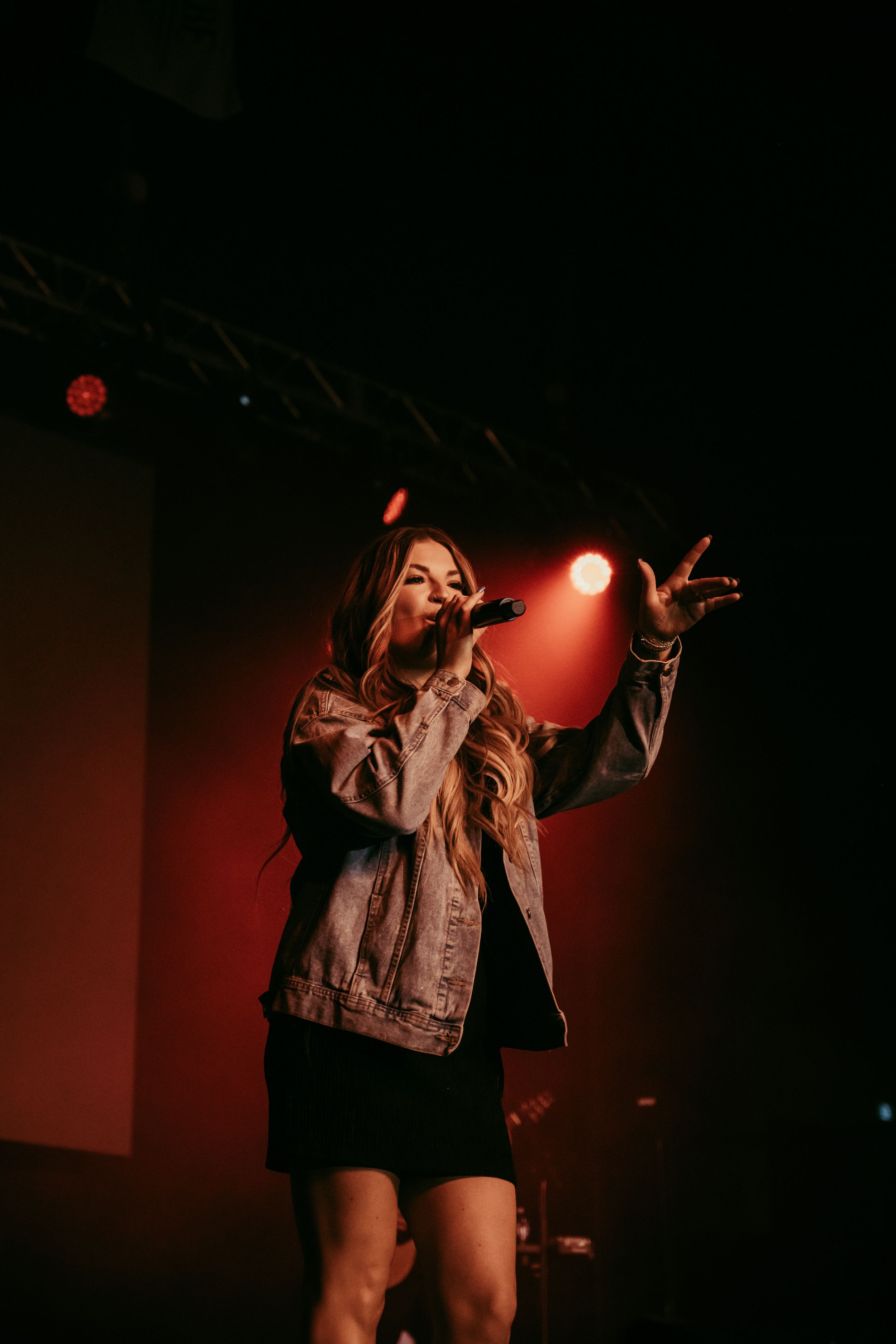 A performer in a denim jacket sings passionately on stage with dramatic red lighting in the background.