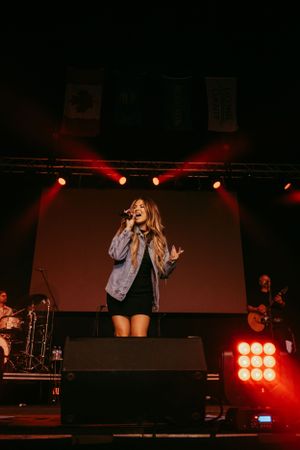 Singer performs on stage in denim jacket and black dress under dramatic red lighting at concert venue.