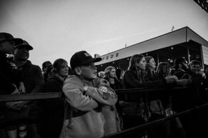 A black and white photograph shows spectators gathered at a railing watching an event outdoors.