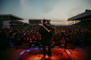 Performer on stage entertaining large crowd at outdoor racetrack venue during sunset.