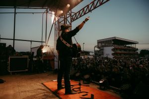 Performer on elevated stage energetically gestures to large crowd at outdoor concert during golden hour.