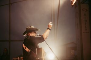 Country music performer in black cowboy hat raising microphone stand on dimly lit concert stage with dramatic lighting effect.