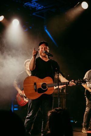 Musician performs with acoustic guitar on smoky stage with spotlights.