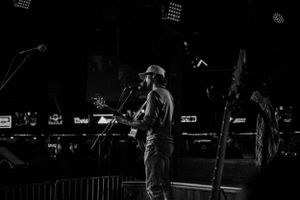 A performer in a baseball cap sings into a microphone on a dimly lit stage during a black and white concert photo.