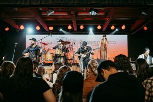 Musicians performing live on stage with instruments at a concert venue while crowd watches in silhouette.