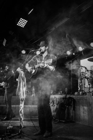 A musician performs on stage with an acoustic guitar amid atmospheric smoke and dramatic concert lighting.