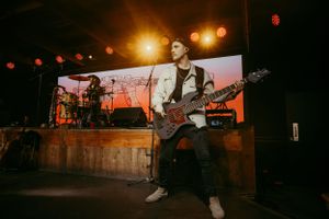 Musician performs energetically on stage with bass guitar against illuminated backdrop during nighttime concert performance.