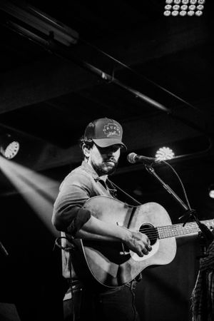 A black and white photo shows a musician performing on stage with an acoustic guitar under dramatic spotlighting.