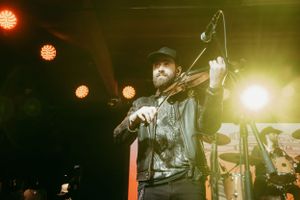 A musician plays violin on stage with dramatic golden lighting and spot lights visible in the background.