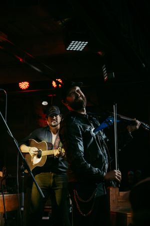 Musicians perform on a dimly lit stage with red lighting, one playing acoustic guitar while another holds a microphone.