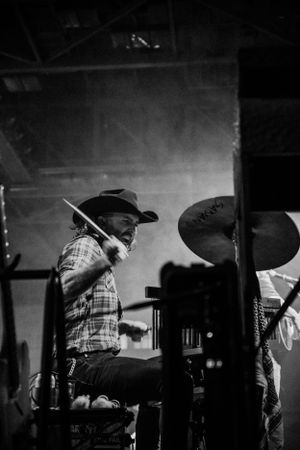 Dramatic black and white photo of a drummer performing on stage wearing a cowboy hat.