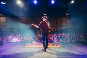 Country music performer plays guitar on stage with crowd visible in background.