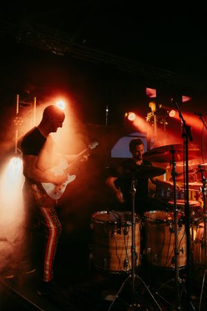 Guitarist silhouetted against orange stage lighting while drummer plays in background.