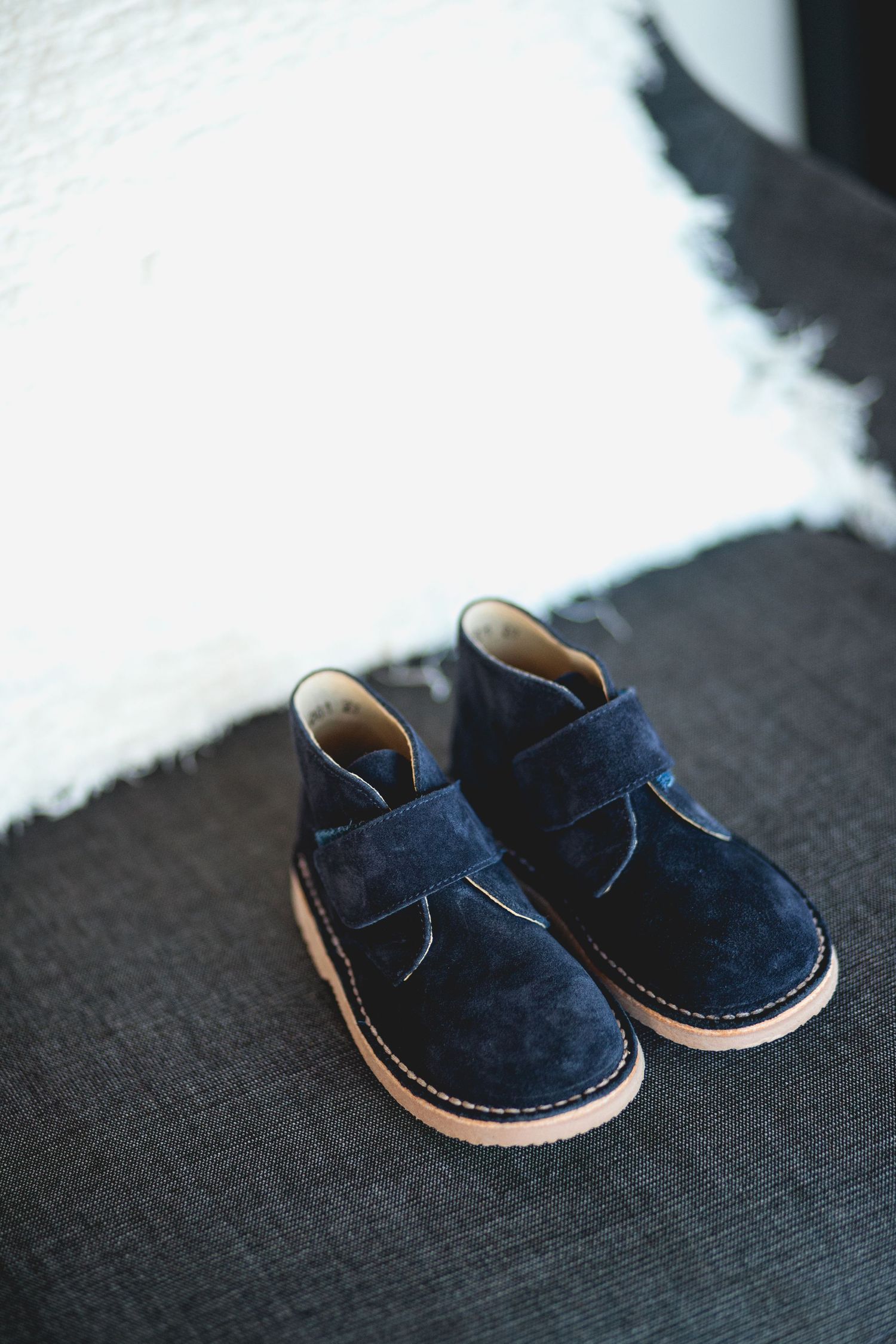 A pair of navy blue suede children's chukka boots with velcro straps sits on a dark gray surface.