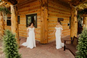 Two brides in white off-shoulder gowns stand before rustic log cabin venue.