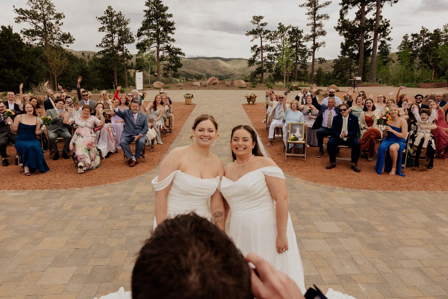 Two brides in white off-shoulder gowns smile at their outdoor wedding ceremony with guests seated behind them.