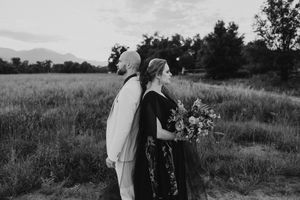 Black and white photo of a couple standing in a field at dusk, woman holding a bouquet, mountains in the background.
