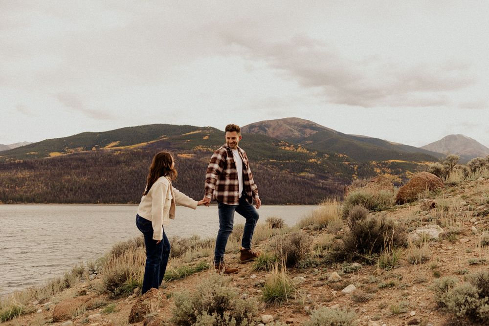 Couple holding hands on a rocky lakeside trail with mountain views and cloudy skies in a scenic outdoor setting.
